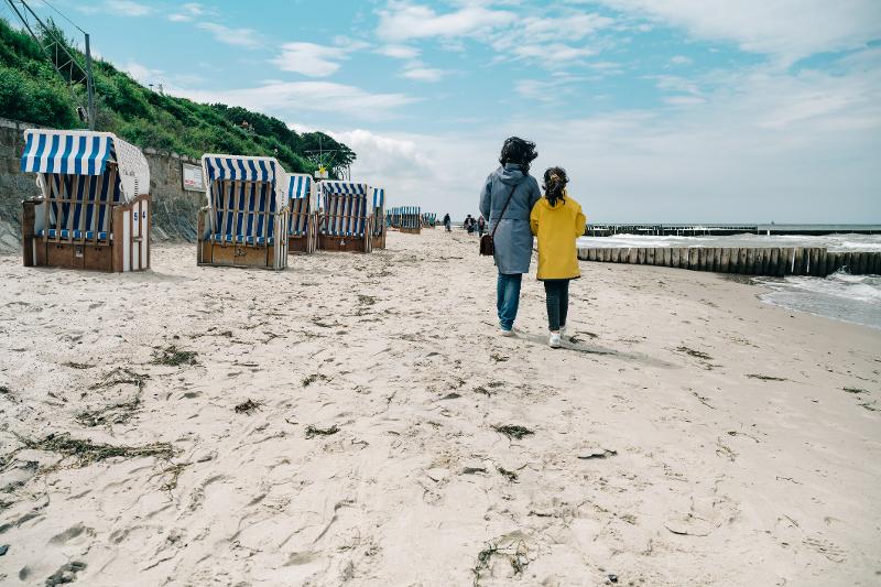 Eine Mutter mit ihrer Tochter, die auf ihrem Familien Kurztrip am Strand entlang gehen. An der Seite sieht man das Meer und auf der anderen Strandkörbe