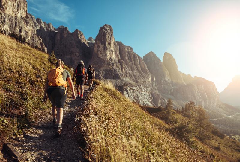 Eine Familie, die auf einem engen Wanderweg läuft, im Hintergrund erkennt man Spitze Berge und Sonnenschein
