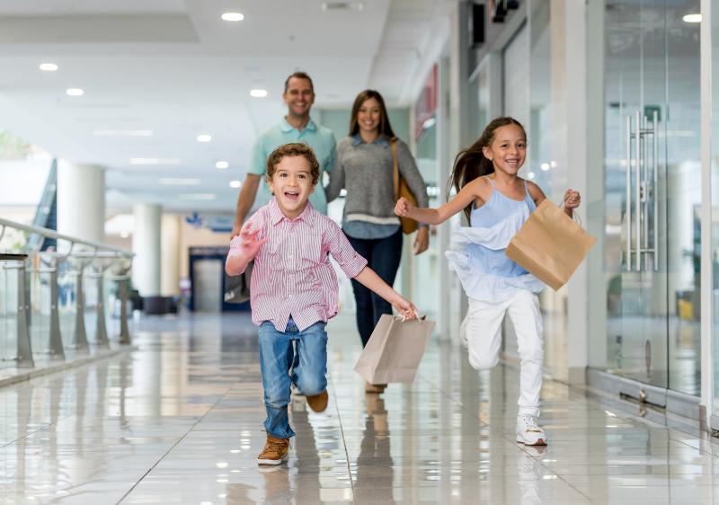 Eine Familie mit zwei Kindern, die im Familien Kurzurlaub Berlin in einer Mall sind. Die beiden Kindern lachen und haben Tüten in der Hand während sie vor den Eltern rennen