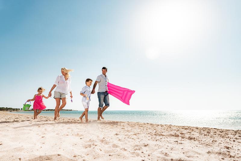 Familie läuft mit Luftmatratze im Gepäck am Strand entlang