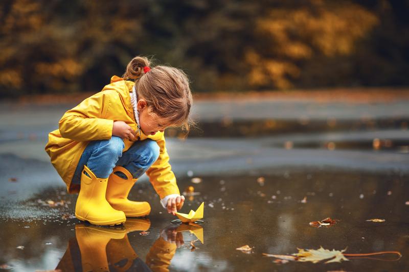 Kind in Regenmantel und Gummistiefeln hockt auf Familienreisen Herbstferien vor einer Pfütze