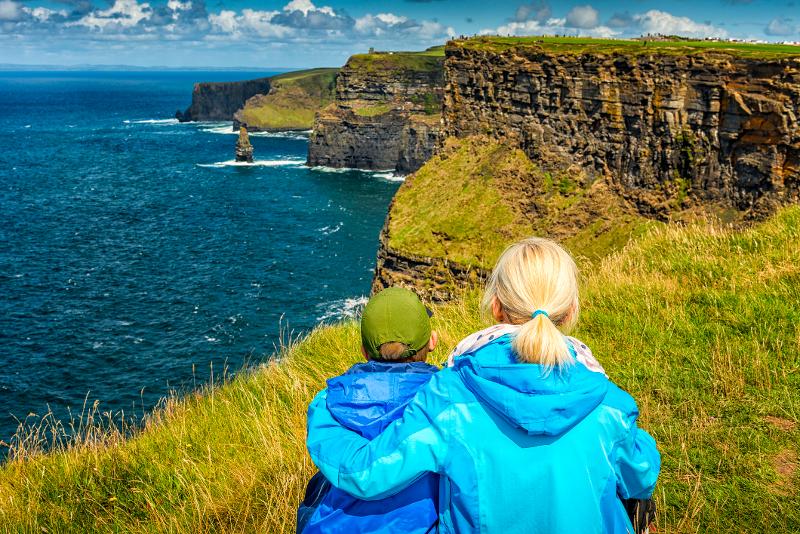 Mutter und Sohn sitzen auf einer grünen Wiese und blicken auf das Meer und die Klippen auf der Familienreise Irland