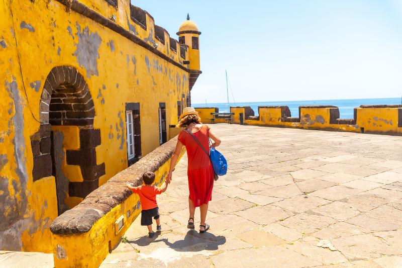 Mutter und Sohn laufen bei schönem Wetter in der Familienreise Portugal neben gelben Gebäude.Im Hintergrund befindet sich das Meer