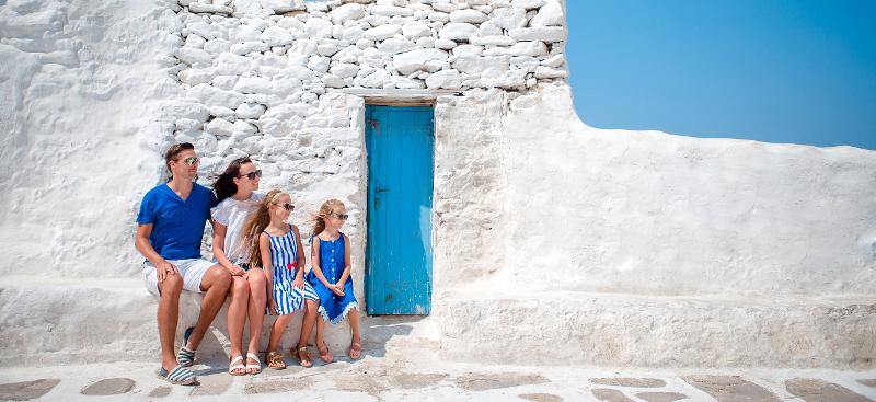 Eine Familie, die im Familienurlaub Athen mit Sonnenbrillen auf einer weißen Steinbank vor einer blauen Tür sitzt