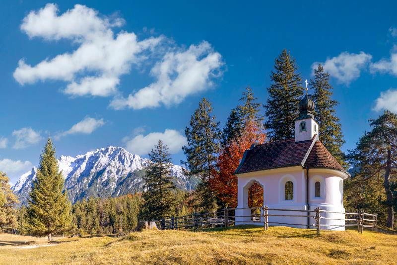 Eine kleine Kirche auf einer Wiese umringt von Bäumen und im Hintergrund sind Schneeberge im Familienurlaub Bayern