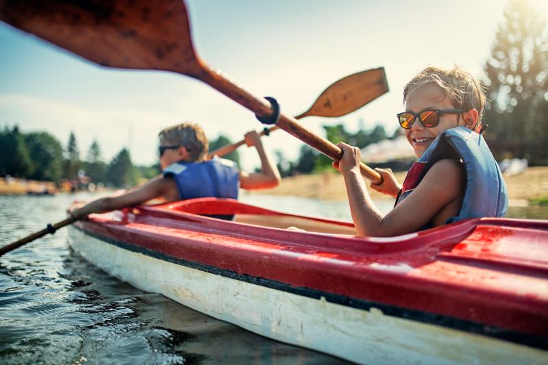 Zwei Kinder, die im Familienurlaub Bayern See mit Schwimmwesten und Sonnenbrillen in einem Kajak auf einem See fahren