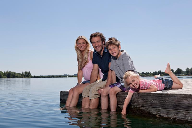 Eine Familie, die im Familienurlaub Bayern See auf einem Steg sitzt und die Füße ins Wasser hält während sie in die Kamera lächeln