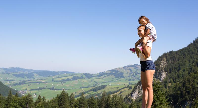 Eine Frau, die ein Kind Huckepack trägt, beide lächeln und im Hintergrund sieht man eine grüne Landschaft mit Häusern im Familienurlaub in den Bergen Deutschland