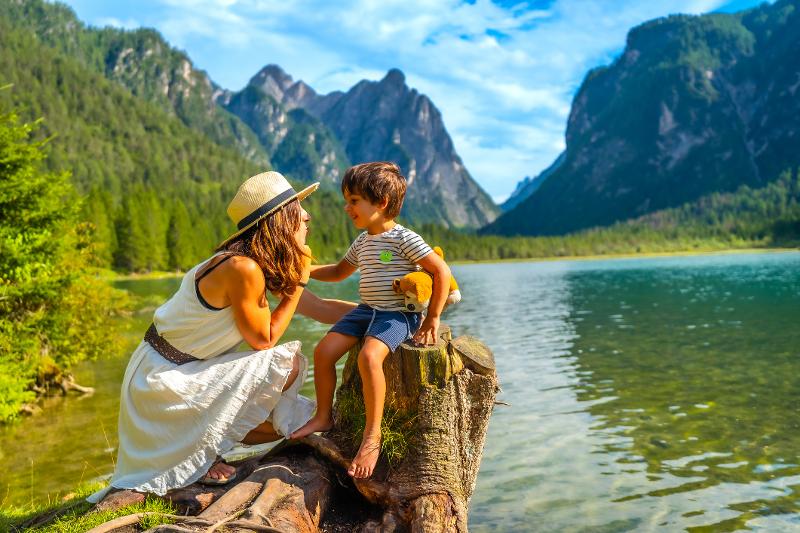  Mutter sitzt mit ihrem kleinen Jungen am See mit bergiger Kulisse im Familienurlaub im Ausland 