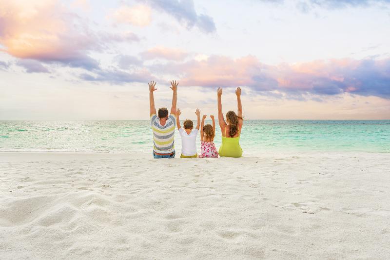 Familie sitzt am Strand und macht die Arme nach oben im Familienurlaub im Oktober am Meer