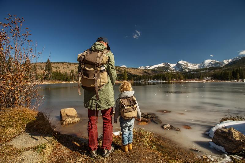 Ein Vater und sein Kind, die im Familienurlaub in den Bergen am See an einem vereisten See mit ihren Rucksäcken stehen. Im Hintergrund sieht man Wälder und Schneeberge