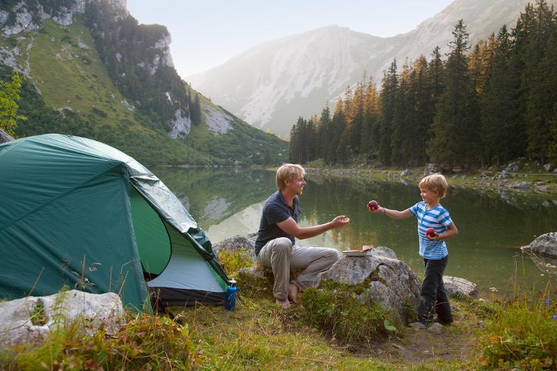 Ein Kind, das seinem Vater einen Apfel reicht. Die beiden sind neben einem Zelt im Familienurlaub in den Bergen am See und im Hintergrund ist ein See und eine bergige Landschaft