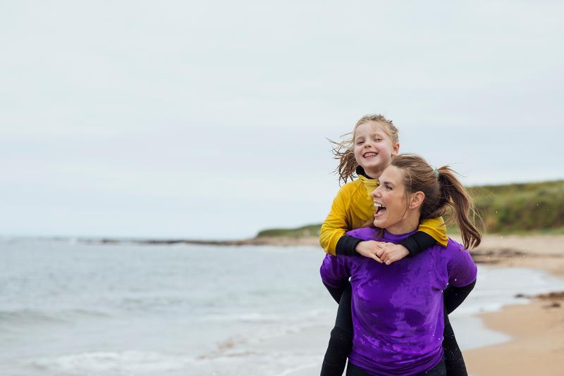  Mutter nimmt Kind am Strand im Familienurlaub in England am Meer Huckepack