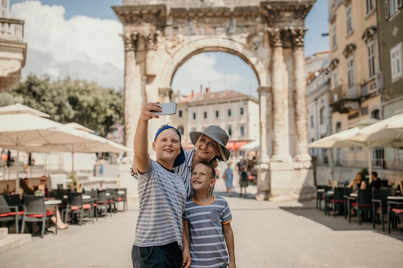Mutter und zwei Kinder machen ein Selfie in einer Stadt. Im Hintergrund ist ein Torbogen und Gebäude, sowie Sonnenschirme und Tische und Stühle eines Restaurants im Familienurlaub Istrien.