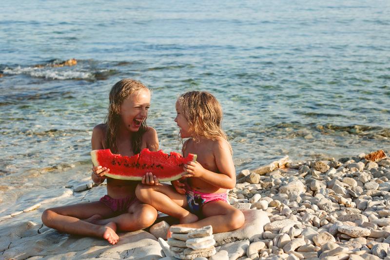 Zwei Kinder sitzen lachend am Strand und essen eine Wassermelone. Im Hintergrund das klare Wasser im Familienurlaub Kroatien Istrien.