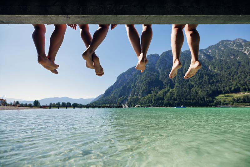 Vier Menschen, die im Familienurlaub Österreich Sommer ihre Füße von einem Steg baumeln lassen, im Hintergrund ein See und Berge