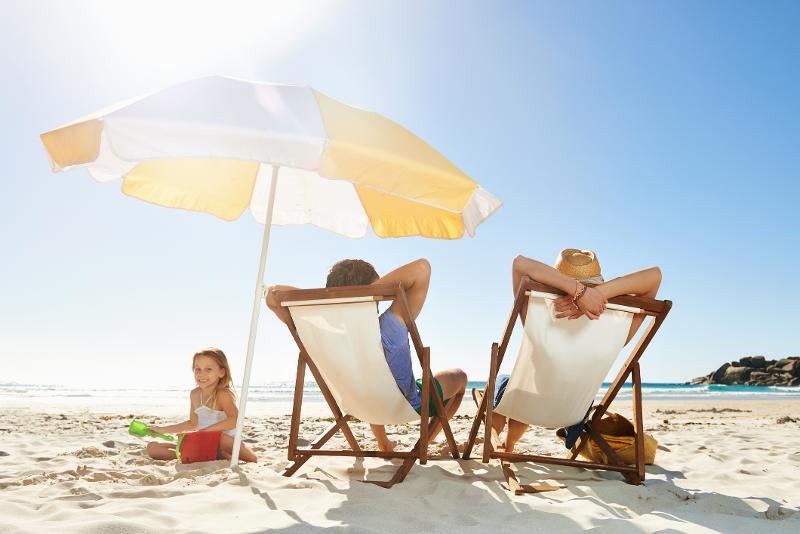 Eltern genießen mit Strandstühlen und Sonnenschirm das Wetter im Familienurlaub Ostsee Last Minute, während ein Kind mit dem Sand spielt 