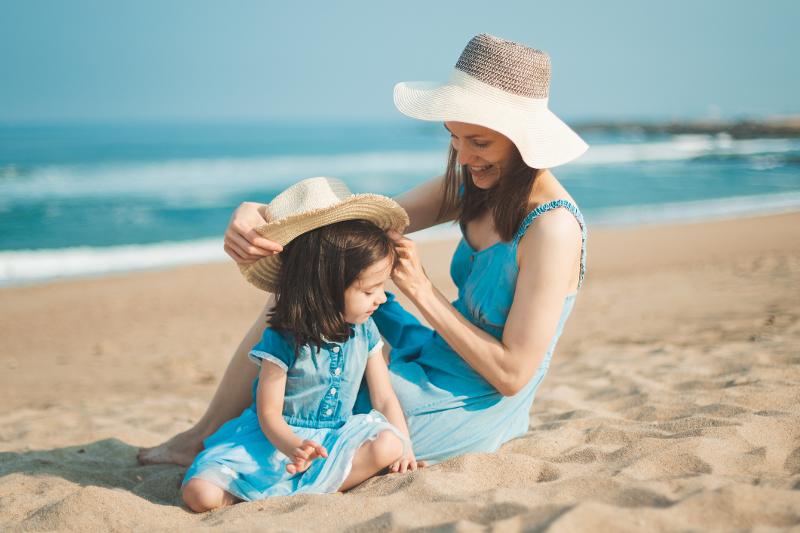 Mutter und Kind mit Hüten, die in blauen Kleiner im Familienurlaub Portugal All Inclusive am Strand sitzen. Im Hintergrund ist das Meer