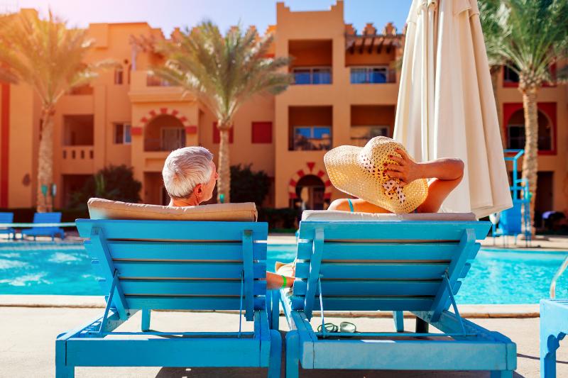 Eine Frau und ein Mann, die im Familienurlaub Portugal All Inclusive auf blauen Sonnenliegen am Pool entspannen. Im Hintergrund sieht man den Pool, Palmen und die orangene Hotelanlage mit Balkonen