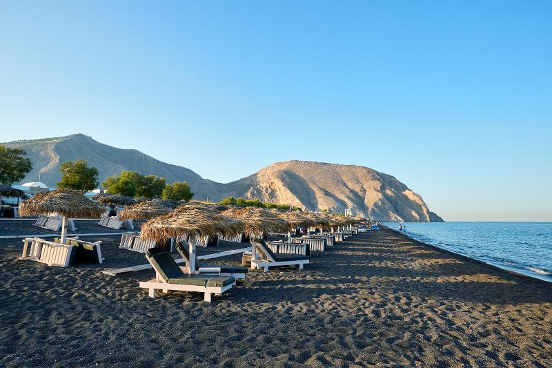 Langer Kiesstrand mit Liegen, Sonnenschirmen, dem Meer und im Hintergrund Berge im Familienurlaub Santorini bei blauem Himmel