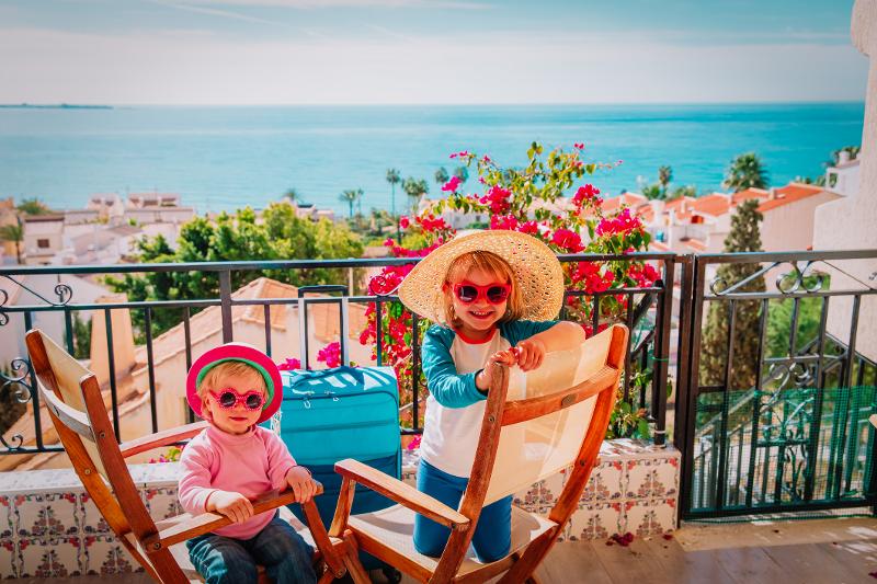 Zwei Kinder, die im Familienurlaub Spanien am Strand in Badebekleidung und Sonnenbrillen auf einem Balkon sitzen und lächeln, im Hintergrund sieht man eine Aussicht auf das Meer