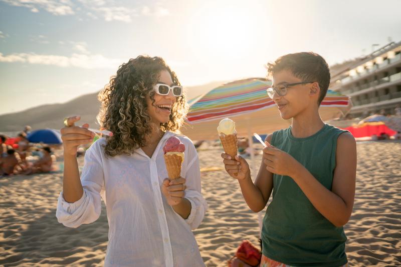 Eine Mutter und ihr Sohn, die im Familienurlaub Spanien am Strand ein Eis in einer Waffel essen und sich grinsend anschauen