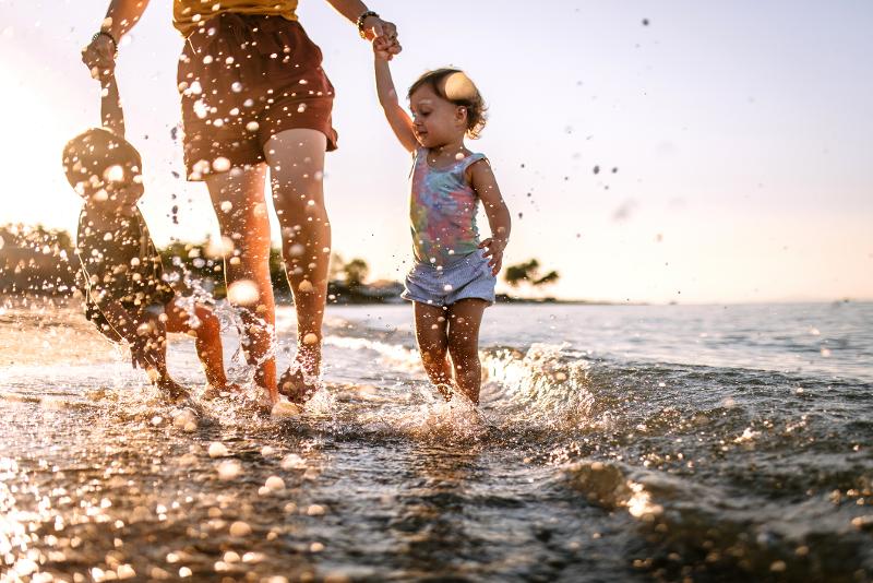 Zwei kleine Kinder, die im Familienurlaub Spanien am Strand die Hände ihrer Mutter halten und im flachen Meer stehen