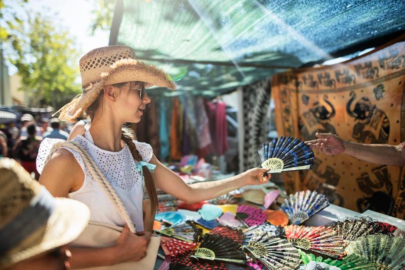 Eine stilvolle Frau mit Strohhut, die bunte Fächer auf einem lebhaften Markt im Familienurlaub Spanien erkundet.