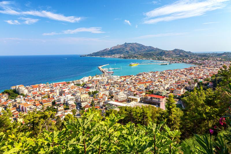 Aussicht auf die Stadt mit Häusern, Steg, Meer und Bergen bei sonnigem Wetter im Familienurlaub Zakynthos