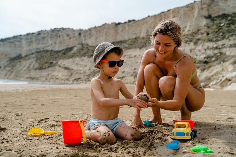 Ein Kind mit Sonnenbrille und Badebekleidung mit seiner Mutter am Strand mit Sandförmchen im Familienurlaub Zypern All Inclusive