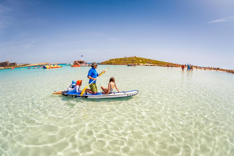Ein Vater, der mit seinen Kindern auf einem Surfboard sitzt und mit einem Paddle im Familienurlaub Zypern All Inclusive durch das flache Meer schwimmt