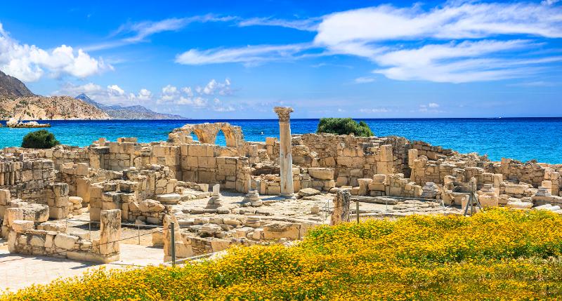 Ein altes Bauwerk mit Säule und zerstörten Steinformationen neben einer Wiese mit gelben Blumen und im Hintergrund das Meer im Familienurlaub Zypern
