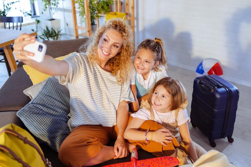 Mutter und zwei Kinder sitzen auf einem Sofa und schießen ein Selfie, im Hintergrund steht ein Koffer
