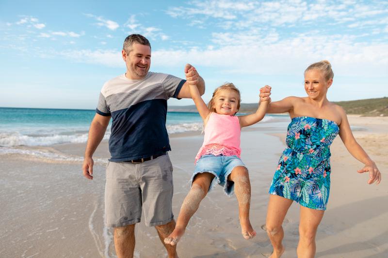Mutter und Vater heben ihre Tochter an ihren Händen hoch, während sie am Strand entlang laufen