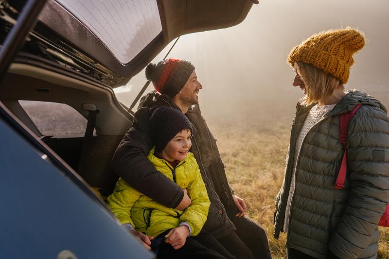 Mutter, Vater und Sohn stehen in herbstlicher Kleidung vorm Auto auf dem Weg in den Herbsturlaub mit Kindern