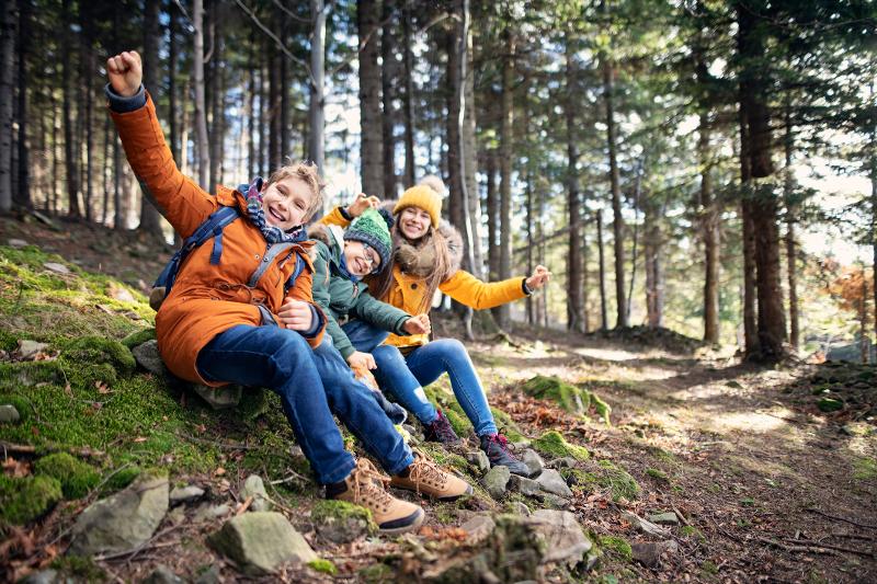 Drei Kinder mit Wanderschuhen sitzen im Herbsturlaub mit Kindern im Wald und haben Spaß