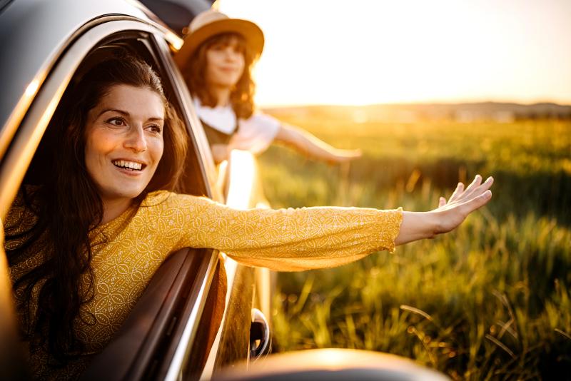 Eine Mutter und ihre Tochter genießen eine Autofahrt bei Sonnenuntergang, beide strecken ihre Hände aus dem Fenster und lächeln.