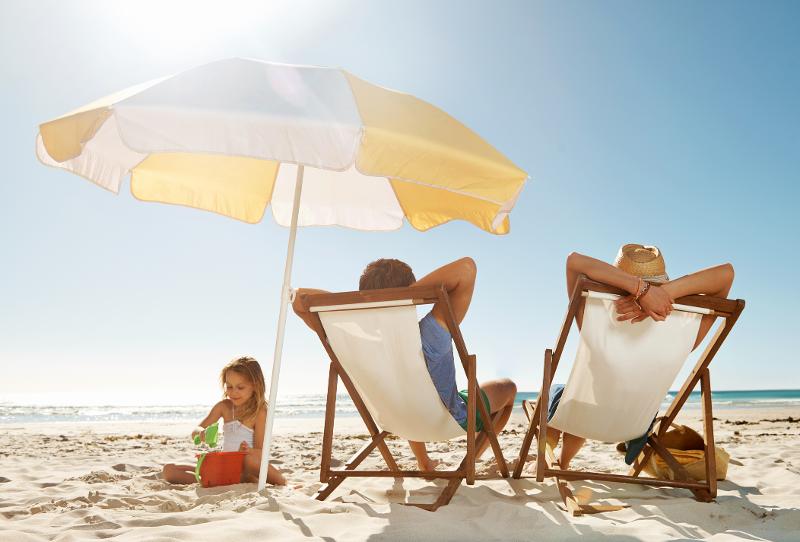 Eltern entspannen auf Liegestühlen unter einem Sonnenschirm, während ihr Kind im Sand spielt, während eines Kinderurlaubs