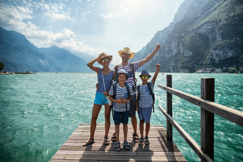 Eine glückliche Familie posiert auf einem Steg am See, umgeben von atemberaubenden Berglandschaften während ihres Kinderurlaubs