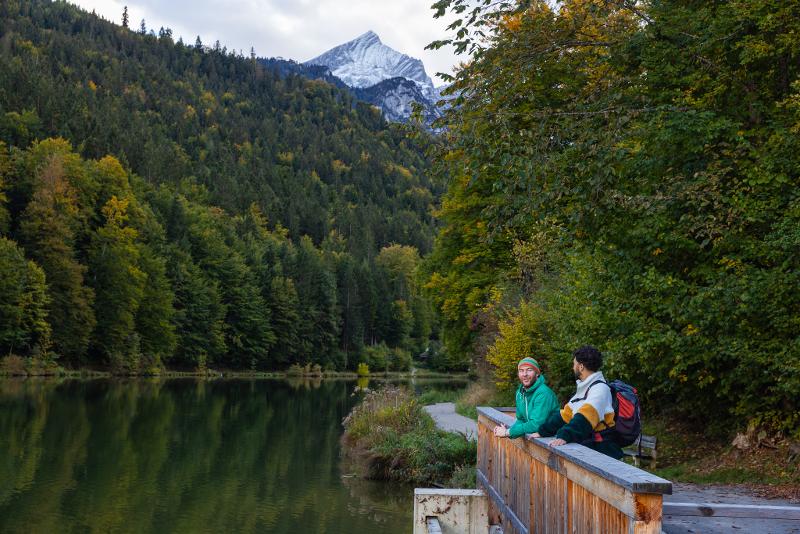 Zwei Männer, die im Kurzurlaub mit Kindern Bayern auf einer Brücke stehen und sie den See anschauen. Im Hintergrund sind Bäume und ein Berg mit Schnee