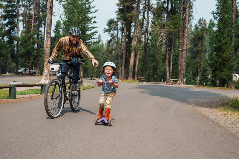 Ein Mann auf einem Fahrrad und sein Kind was auf einem Roller im Kurzurlaub mit Kindern vor ihm fährt