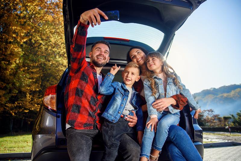 Eine Familie, die im Kurzurlaub mit Kindern Niedersachsen in ihrem Kofferraum sitzt und ein Selfie von sich macht. Im Hintergrund sieht man blauen Himmel und Wälder