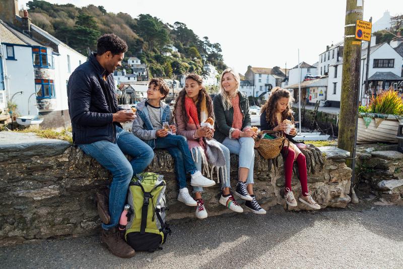 Eine Familie mit drei Kindern, die im Kurzurlaub mit Kindern auf einer Steinmauer sitzen und etwas essen. Im Hintergrund sieht ma Häuser, eine Straße und Bäume