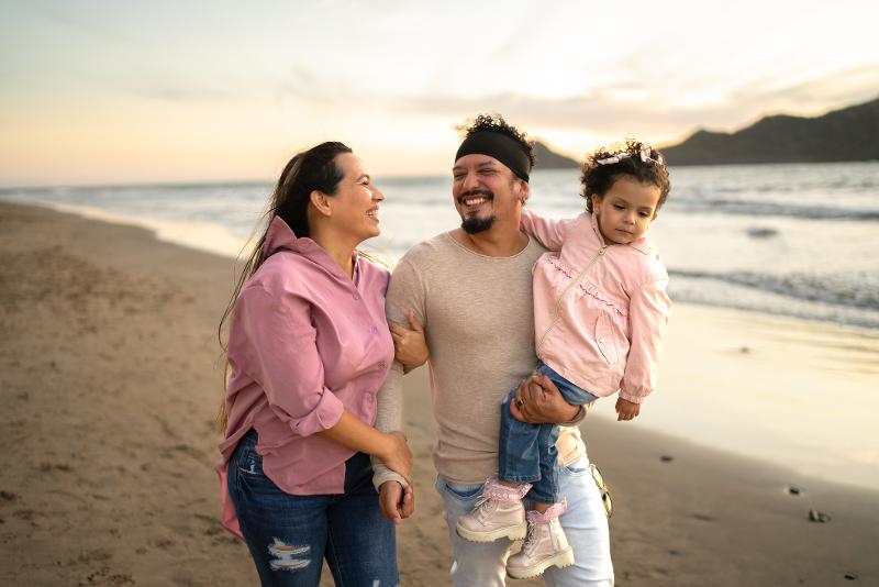 Eine Familie mit kleinem Kind, die im Kurzurlaub mit Kindern einen Strandspaziergang unternimmt, im Hintergrund das Meer und Berge bei Dämmerung