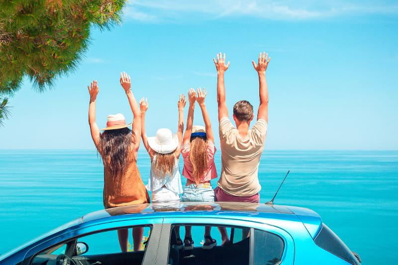 Eine paar Eltern sitzen mit ihren zwei Kindern auf dem Dach ihres Autos und blicken auf das blaue Meer im Last Minute Familienurlaub Italien