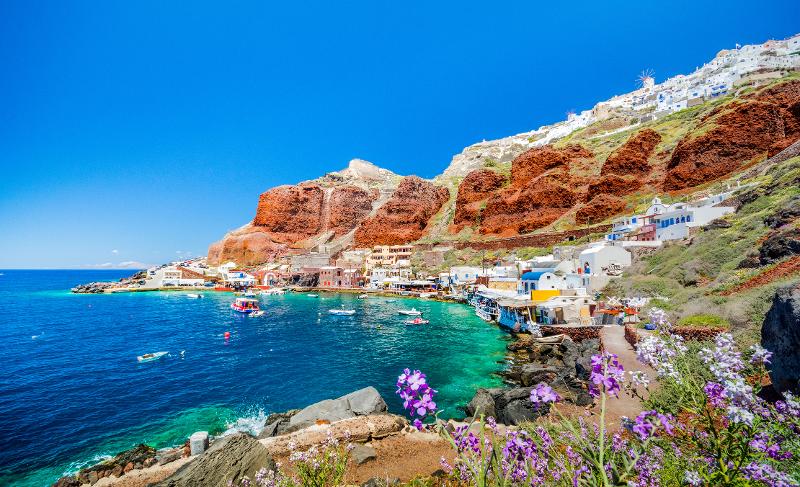 Aussicht auf eine Kleine Ortschaft mit Häusern, dem Meer mit Booten und Felsen im Hintergrund im Santorini Urlaub mit Kindern