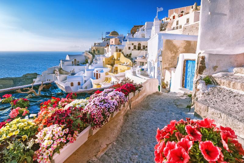 Eine schmale Straße mit Blumen, weißen Häusern und dem Meer am Horizont im Santorini Urlaub mit Kindern