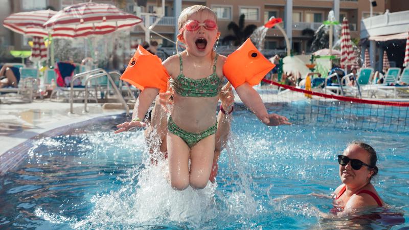 Ein Kind in Badebekleidung das im Spanien Urlaub mit Kindern All Inclusive in einem Pool hoch ins Wasser geworfen wird