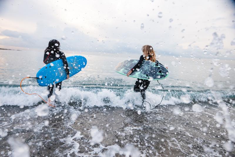 Zwei Kinder, die im Neoprenanzug mit Surfboards im Spanien Urlaub mit Kindern am Meer ins Wasser rennen. Es ist bewölkt und die Tropfen des Meeres umranden das Meer