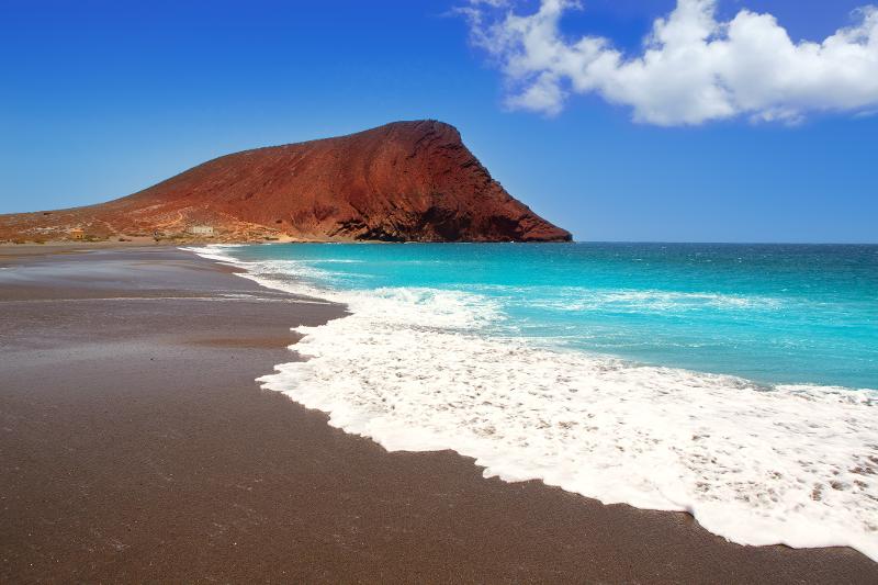 Ein Strandabschnitt mit dunklem Sand und türkisem Meer im Spanien Urlaub mit Kindern am Meer. Im Hintergrund ein großer Hügel bei schönem Wetter.